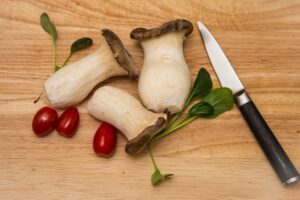 Three fresh cardoncelli mushrooms on a wooden chopping board, with cherry tomatoes and a knife