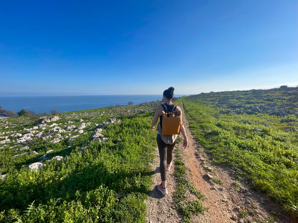 A walker with a backpack on a dirt track through green, stony countryside above the sea in the Salento