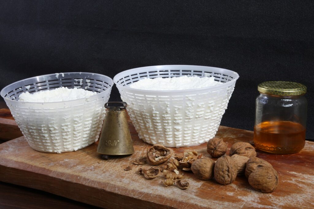 Fresh ricotta in two draining baskets on a wooden board, with walnuts, honey and an old cowbell