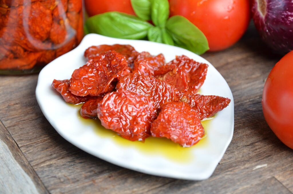 Sun-dried tomatoes in olive oil on a white dish, with fresh tomatoes, basil and a jar of preserved tomatoes behind